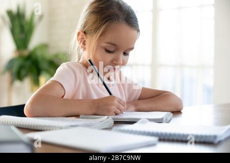 Happy little schoolgirl sitting at table, doing homework. Stock Photo