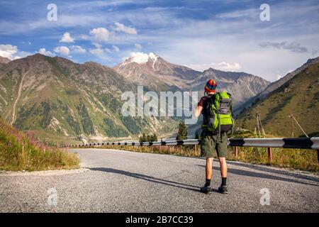 Tourist with backpack and rainbow hat walking down the road in the mountains near Big Almaty Lake in Kazakhstan Stock Photo
