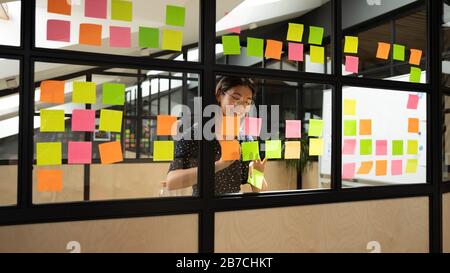 Excited Asian businesswoman wearing glasses writing notes on stickers Stock Photo