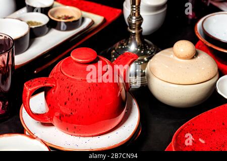 Red porcelain shiny teapot with black dots on a black table among other tea utensils, plates and handmade containers. Selective focus. Stock Photo