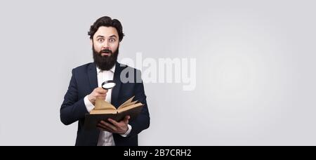 shocked bearded man reading book with magnifier, male over gray background, panoramic mock-up with space for text Stock Photo