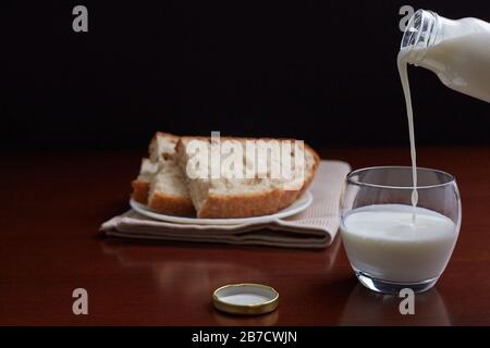 Kefir from a bottle flows into a glass. Bread in the background. Dark background and brown wooden table. Stock Photo
