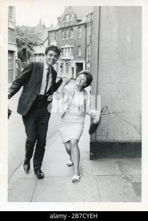 Young smartly dressed good looking 1960s couple in love skipping down a street holding hands with Georgian buildings in background. Original 1960s vintage snapshot vernacular photography found photo UK social history. Photographer unknown (Richard Bradley collection) Stock Photo