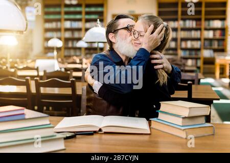 Handsome senior bearded man grandfather hugging and kissing his cute granddaughter, little girl in eyeglasses, sitting at the table with many books in Stock Photo