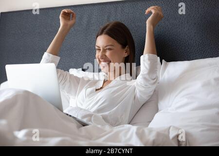 Emotional overjoyed young woman celebrate success, sitting in bed. Stock Photo