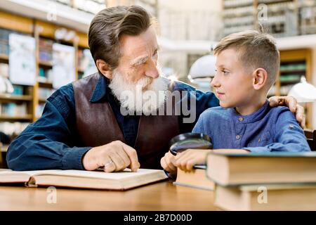 Happy smiling little boy with his cheerful bearded grandfather reading books at library, looking each other. Smiling little boy with his senior teache Stock Photo