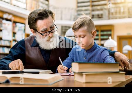 Senior man teacher or grandfather teaching little boy, his grandchild, reading book in library, while concentrated boy makes notes in his copy book. E Stock Photo