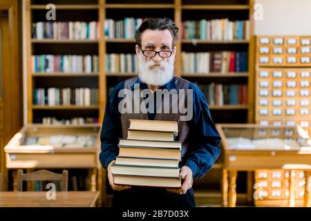 Portrait of attractive bearded old man wearing shirt and leather vest, high school teacher or librarian, holding books in hands, standing in vintage l Stock Photo