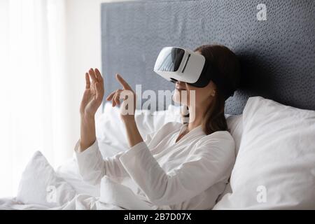 Happy young brunette woman resting in bed, wearing VR googles. Stock Photo