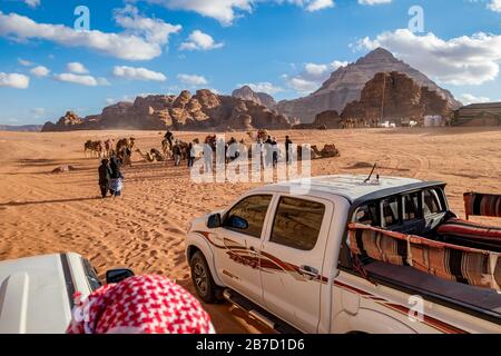 WADI RUM, JORDAN - JANUARY 31, 2020: Tourists meet locals and camels for a ride, view from safari SUV top on red sand. Winter puffy clouds windy afternoon sky, desert, Hashemite Kingdom of Jordan Stock Photo