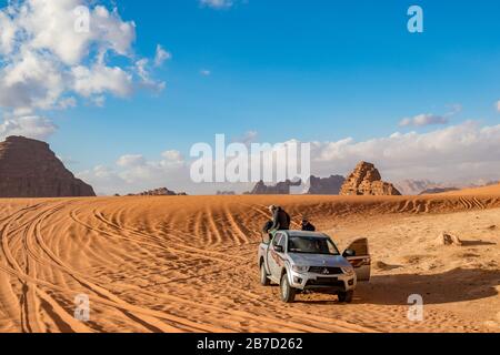 WADI RUM, JORDAN - JANUARY 31, 2020: Few people prepare for a ride on safari SUV for tourists on the red sand. Winter puffy clouds windy afternoon sky. beautiful desert, Hashemite Kingdom of Jordan Stock Photo