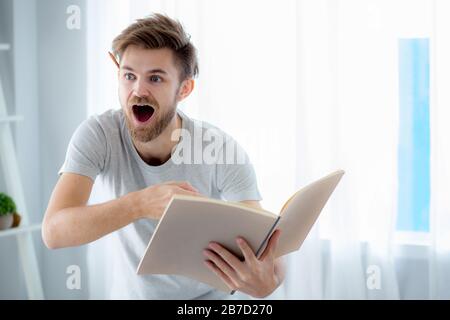 Handsome guy in eyeglasses is reading book preparing exam with standing at the living room - education concept. Stock Photo