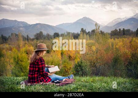 Woman in red checked shirt and hat sitting on the grass reading book at autumn forest and mountains background Stock Photo