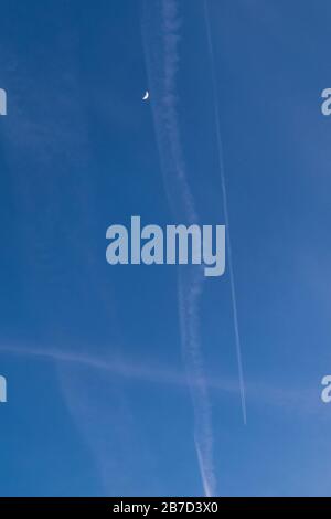 Crossing of airplane trails with crescent moon in blue sky Stock Photo