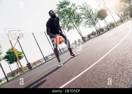Young bearded african man wearing virtual reality headset playing basketball on court outdoors concentrated watching video Stock Photo