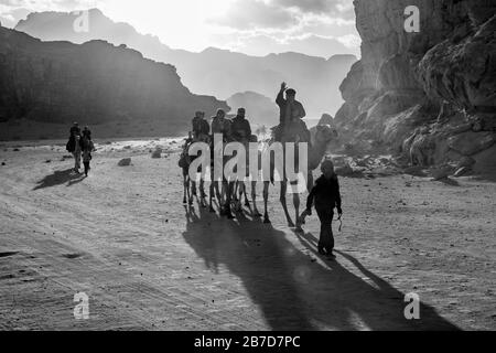 WADI RUM, JORDAN - JANUARY 31, 2020: Impressive sky with sun rays over people riding camels. Black and white image, winter puffy clouds afternoon sky. Desert, Hashemite Kingdom of Jordan Stock Photo