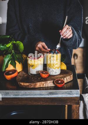 Woman eating overnight oat bircher muesli with mango smoothie, buckwheat granola and fresh berries from glass with spoon over grey concrete kitchen co Stock Photo