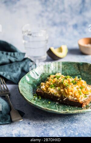 Breakfast. Scrambled eggs on rye bread with chive with avocado in th ebackground and glass of water. On blue background with chive with avocado in th Stock Photo