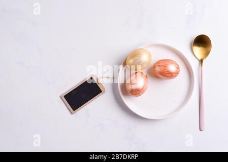 Happy Easter card with handmade painted eggs on a ceramic plate and golden spoon on a light grey marble background, copy space. Easter concept. Stock Photo