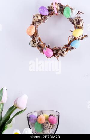 Easter card with handmade wreath from willow branches and painted colorful eggs, tulips flowers in vase against light grey wall. Stock Photo