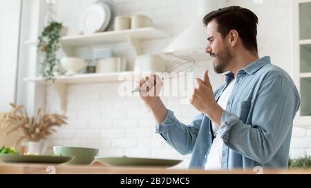 Funny handsome man singing into whist, having fun in kitchen Stock Photo