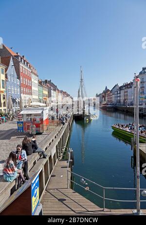 View along the Nyhavn canal on a sunny and nearly warm spring day. Copenhagen, Denmark. Copenhageners and tourists enjoy the sunshine. Stock Photo