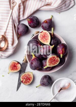 A bowl of ripe, purple figs on a white marble countertop surrounded by a striped napkin, rose gold spoons, and a small bowl of sugar Stock Photo