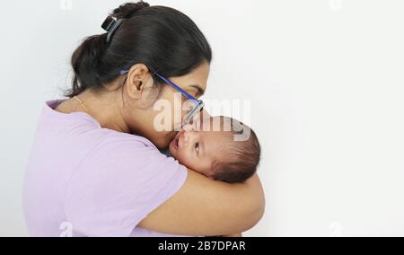 a young mother in spectacles kissing her infant baby boy in her lap in solid grey background with space for text. mothers day concept photo. Stock Photo