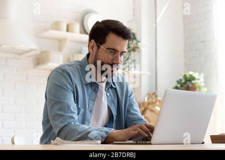 Focused young man wearing glasses using laptop, typing on keyboard Stock Photo