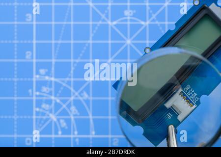 Close-up of circuit board with lcd display held by crocodile-clips seen through magnifying glass on blue background. Stock Photo