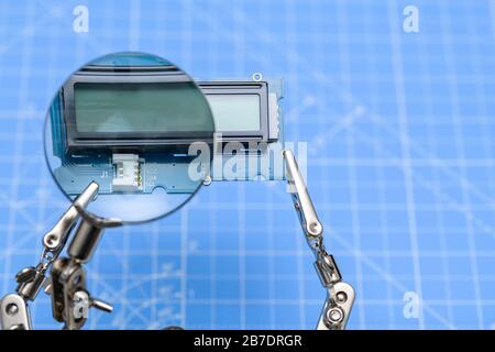 Close-up of circuit board with lcd display held by crocodile-clips seen through magnifying glass on blue background. Stock Photo