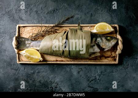 Cooked fish sea bass baked in bamboo leaves with lemon and rosemary on rectangular ceramic plate over dark texture concrete background. Flat lay, spac Stock Photo