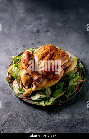 Whole baked chicken poultry on ceramic plate, served with green beetroot salad leaves on linen cloth over black concrete background. Copy space. Dinne Stock Photo