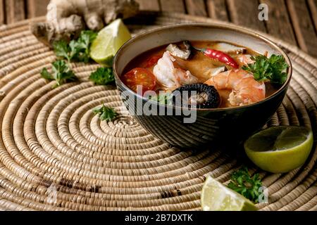 Traditional spicy Thai soup tom yum kung with shiitake mushrooms and prawns, ingredients above on straw wicker napkin over wooden plank table. Stock Photo