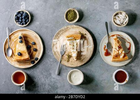 Pieces of cheesecake with different toppings on ceramic plates and variety of sauces in ceramic bowls in row over grey texture background. Flat lay, s Stock Photo