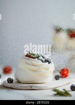 Icing sugar sprinkles on mini Pavlova cake with fresh berries and fresh green rosemary. Vertical composition with white marble tabletop and white wall Stock Photo