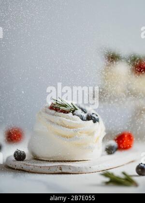 Icing sugar sprinkles on mini Pavlova cake with fresh berries and fresh green rosemary. Vertical composition with white marble tabletop and white wall Stock Photo