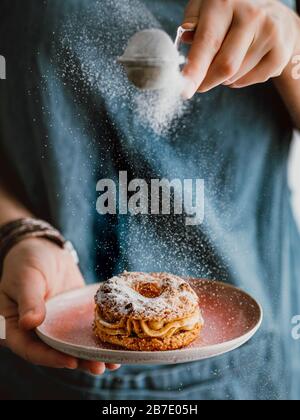 Woman in blue denim apron holds with one hand pink plate with choux au craquelin cake and sprinkles with her other hand icing sugar through a sieve's. Stock Photo