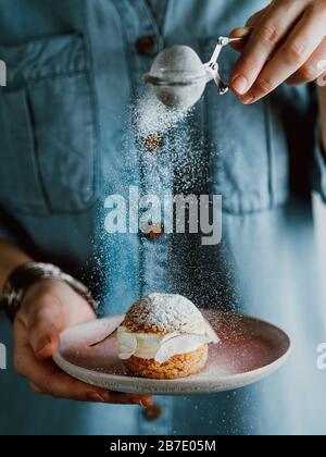 Woman in blue denim shirt holds with one hand pink plate with choux au craquelin pastry and sprinkles with her other hand icing sugar through a sieve' Stock Photo