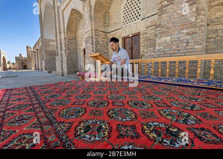 Uzbek man sitting on the carpet and reading in the courtyard of Chor Bakr religious complex, in Bukhara, Uzbekistan Stock Photo
