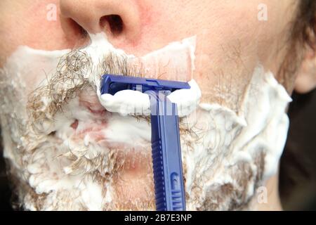 A man shaves his beard with a blue disposable safety razor. White shaving foam on the cheeks and chin. Stock Photo