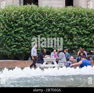 City workers eat lunch outside & chat next to the fountain at Cabot Square, Canary Wharf, London, on a hot July day. Stock Photo