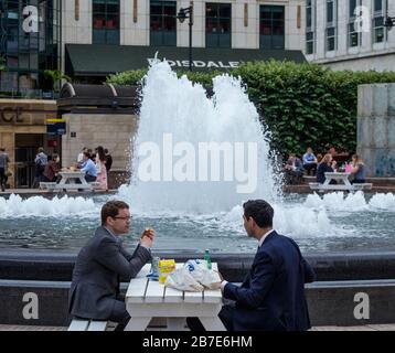 Two men in suits each and chat at outside table next to water fountain during lunchtime at Cabot Square, Canary Wharf, London, on a warm day. Stock Photo