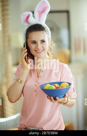 happy modern 40 years old woman in a pink blouse and easter bunny ears with a blue plate with colorful easter eggs talking on a mobile phone in the mo Stock Photo