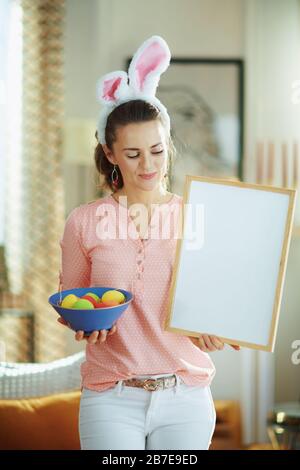 smiling elegant woman in a pink blouse and easter bunny ears with a blue plate with colorful easter eggs showing blank white billboard in the modern h Stock Photo