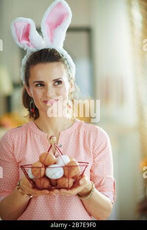 pensive young woman in a pink blouse and easter bunny ears with basket with eggs for coloring looking at copy space at modern home in sunny spring day Stock Photo