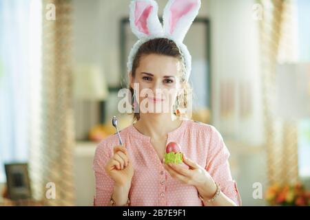 Portrait of 40 years old woman in a pink blouse and easter bunny ears with spoon and red easter egg in green egg cup in the modern house in sunny spri Stock Photo