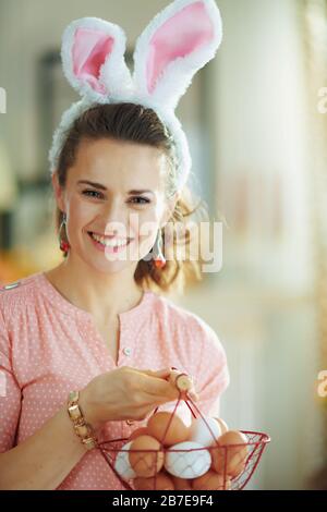 smiling elegant female in a pink blouse and easter bunny ears holding basket with eggs for coloring in the modern living room in sunny spring day. Stock Photo