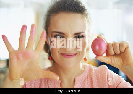 concerned modern housewife in a pink blouse and easter bunny ears showing hands stained with a red easter egg in the modern house in sunny spring day. Stock Photo