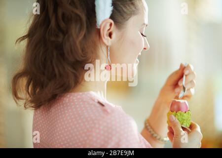 Seen from behind young female in a pink blouse and easter bunny ears eating red easter egg in green egg cup with spoon at modern home in sunny spring Stock Photo